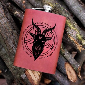 Puede incluir: Una petaca de cuero roja con un diseño grabado en negro de una cabeza de cabra con cuernos dentro de un pentagrama. El texto alrededor del diseño dice "Omnium Templum Pacis Albus 666 Templi Omnium Templum Pacis Albus 666".