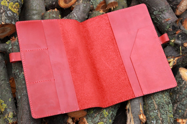 Puede incluir: Carpeta de cuero roja para cuaderno con varios bolsillos y cierre de solapa. La carpeta est&aacute; abierta para revelar un interior de ante rojo.