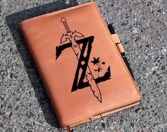 May include: A brown leather journal with a black design of a sword and a flower. The design is a stylized letter Z with a sword going through it. The journal has a pen holder and a pen.