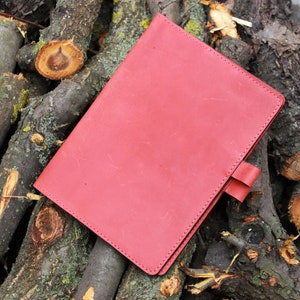 Puede incluir: Un cuaderno de cuero rojo con un borde cosido y un cierre de solapa. El cuaderno est&aacute; sobre una pila de ramas de &aacute;rbol.