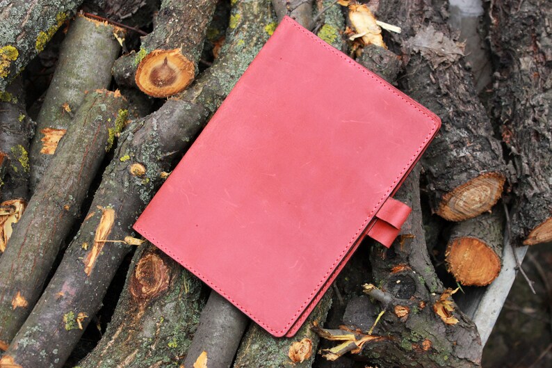 Puede incluir: Un cuaderno de cuero rojo con un borde cosido y un cierre de solapa. El cuaderno est&aacute; sobre una pila de ramas de &aacute;rbol.