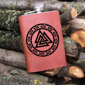 Puede incluir: Una petaca de cuero marrón con una tapa de acero inoxidable y un diseño grabado en negro de un nudo celta con un triángulo en su interior.