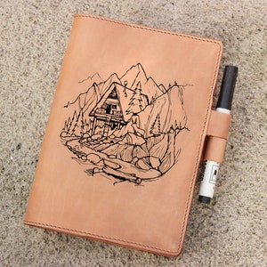 May include: A brown leather journal with a black pen tucked into the side pocket. The cover features an engraved design of a cabin in the mountains with a river flowing through the scene.