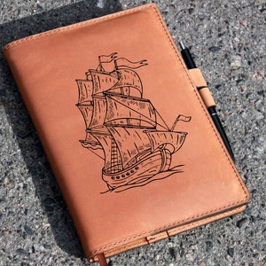 May include: A brown leather journal with a pen holder and an engraved image of a sailing ship.