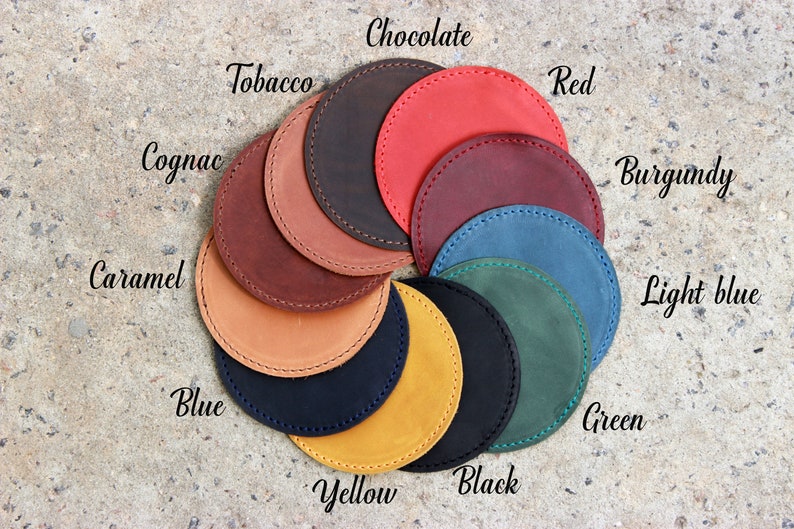 Puede incluir: Una variedad de posavasos de cuero en diferentes colores, incluyendo negro, marr&oacute;n, rojo, azul, verde, amarillo y caramelo. Los posavasos est&aacute;n dispuestos en un c&iacute;rculo con los nombres de los colores escritos encima de cada posavasos.