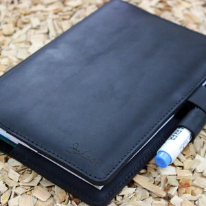 Puede incluir: Una libreta de cuero negro con un portaplumas y un bol&iacute;grafo insertado. La libreta tiene un borde cosido y un peque&ntilde;o logotipo en la parte delantera.