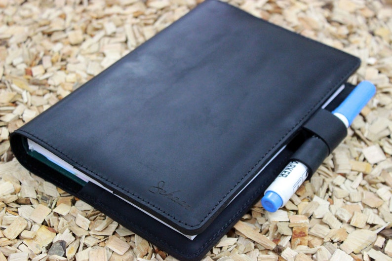 Puede incluir: Una libreta de cuero negro con un portaplumas y un bol&iacute;grafo insertado. La libreta tiene un borde cosido y un peque&ntilde;o logotipo en la parte delantera.