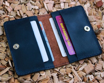 May include: A blue leather wallet with a brown leather interior and a snap closure. The wallet is open and has several cards inside, including one with the text "ВЫПРАЮТЬ УСІ!" in pink.