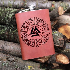 Puede incluir: Una petaca de cuero marrón con una tapa plateada y un diseño en blanco y negro de un símbolo triquetra dentro de un círculo con líneas radiantes.