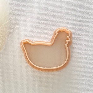 May include: A light brown plastic chicken cookie cutter. The cutter is shaped like a chicken with a rounded body and a small head with a comb and wattle.