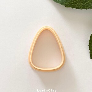 May include: A light brown plastic cookie cutter shaped like a triangle with rounded edges. The cutter has a textured edge.