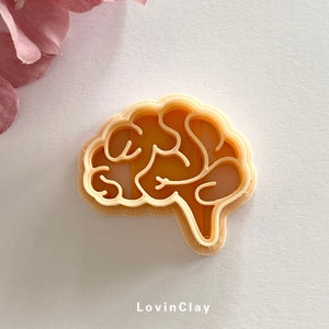 May include: A light brown plastic cookie cutter shaped like a brain. The cutter has a double layer design, with a raised outline and a cut-out inside.