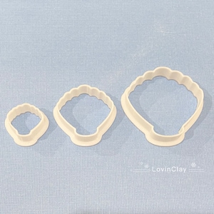 May include: Three white plastic cookie cutters shaped like seashells. The cutters are different sizes, with the largest cutter at the top of the image and the smallest cutter at the bottom.