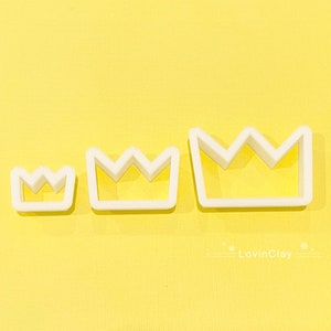 May include: Three white plastic crown-shaped cookie cutters of different sizes on a yellow background. The largest cutter is at the top, the smallest at the bottom.
