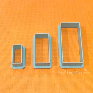 May include: Three blue plastic rectangle cookie cutters of different sizes. The largest cutter is about 5 cm wide and 7.5 cm long. The smallest cutter is about 2.5 cm wide and 3.75 cm long.