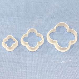 May include: Three white plastic cookie cutters shaped like four-leaf clovers. The cutters are different sizes, with the largest cutter at the top of the image and the smallest cutter at the bottom.