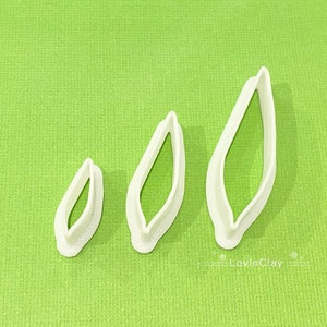 May include: Three white plastic leaf-shaped cookie cutters of different sizes. The cutters are on a green surface.