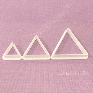 May include: Three white plastic triangle cookie cutters of different sizes. The cutters are on a purple textured surface.