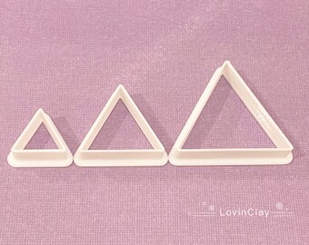 May include: Three white plastic triangle cookie cutters of different sizes. The cutters are on a purple textured surface.