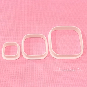 May include: Three white plastic square cookie cutters of different sizes on a pink background. The largest cutter is at the back, the medium cutter is in the middle, and the smallest cutter is at the front.