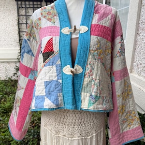 May include: A cropped jacket constructed from a patchwork quilt featuring various floral and geometric patterns in shades of pink, blue, and white. The garment includes a bright blue trim, long sleeves, and two toggle closures with white leather-like tabs.