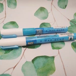 Könnte beinhalten: Zwei blaue und weiße Zig 2-Wege-Klebestifte. Die Stifte sind säurefrei und xylenfrei. Der Text auf den Stiften lautet "Säurefrei - Xylol-frei 2-Wege-Kleber".