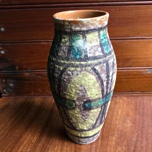 May include: A ceramic vase with a textured, matte finish featuring a geometric pattern in shades of yellow, brown, and teal. The vessel has a cylindrical shape with a slightly flared rim and a rough, earthy surface texture throughout the exterior.