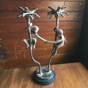 May include: A bronze-toned candle holder featuring two monkeys holding hands, climbing palm tree-shaped candle holders. The base is a dark, circular shape. The background is a wooden surface.