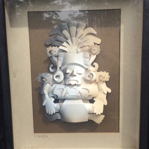 Puede incluir: Una escultura de papel enmarcada de una figura blanca estilizada con un tocado. La figura tiene una cara grande y redonda con una boca ancha y los ojos cerrados. La escultura está sobre un fondo marrón y firmada "EISNER" en la esquina inferior izquierda.