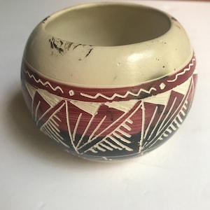 Op de afbeelding: Een kleine, ronde, crèmekleurige keramische kom met een brede band van donkerrode en zwarte geometrische patronen. Het ontwerp bevat gegraveerde witte lijnen die driehoeken en zigzagpatronen rond het midden vormen, met een gladde, matte afwerking.