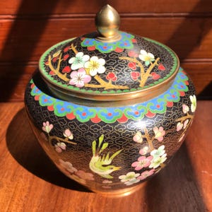 May include: A round black cloisonné jar with a matching lid featuring a gold-toned finial. The surface is decorated with colourful floral patterns, including white and pink blossoms on gold branches, and a small green bird, all set against a black background with fine gold wire detailing.