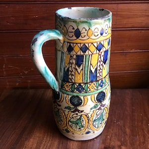 May include: A ceramic pitcher with a blue, green, yellow, and white geometric pattern. The pitcher has a handle and a rounded body.