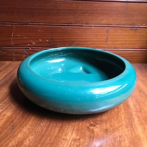 May include: A round, teal-colored ceramic bowl with a smooth, glossy finish. The bowl features a wide, thick rim and a shallow, concave interior basin. The design is simple and circular, showcasing a vibrant, solid green-blue glaze throughout.