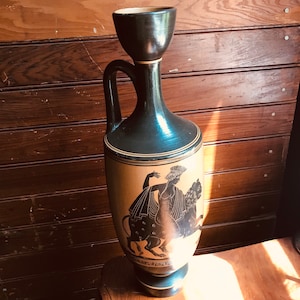 May include: A black and gold ceramic vase with a black silhouette of a woman riding a mythical creature. The vase has a wide mouth and a narrow neck.