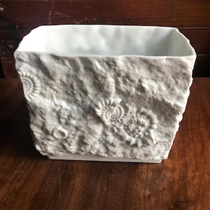 May include: White ceramic square vase with a textured, stone-like exterior. The surface is embossed with fossil designs. The interior is smooth and white. The vase sits on a wooden surface.
