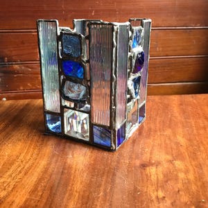 May include: A square glass candle holder with a geometric design. The holder is made of clear and blue glass and has a silver metal frame.