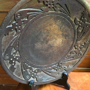 May include: A dark brown wooden plate with a carved design of birds and flowers around the edge. The plate is on a black stand.