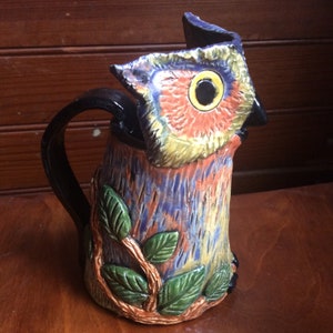 Peut inclure: Une cruche en céramique en forme de hibou avec une poignée noire. Le hibou est peint dans des tons de bleu, vert, orange et jaune. Les yeux du hibou sont noirs avec des centres jaunes. La cruche a un motif de feuilles autour du bas.