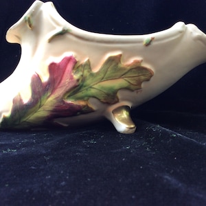 May include: A white ceramic bowl with a gold trim and an oak leaf design in shades of green, brown, and pink. The bowl is shaped like a cornucopia and has three feet.
