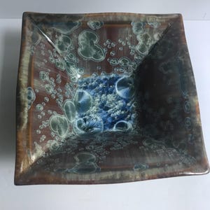 May include: A square ceramic bowl featuring a crystalline glaze with a deep brown base color. The interior surface displays a pattern of light blue and white circular crystal formations, with a concentrated cluster of blue starburst patterns at the center.