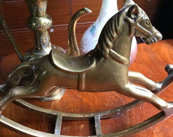 May include: A brass rocking horse figurine with a saddle and stirrups. The horse is on a wooden surface with a brass teapot and vase in the background.
