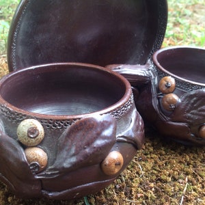 Puede incluir: Dos tazas de cerámica marrón con diseños de hojas y bayas. Las tazas tienen un aspecto rústico y hecho a mano y son perfectas para servir bebidas calientes.