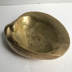 May include: A gold-colored metal dish shaped like a clam shell. The interior surface has a textured, uneven finish with a metallic sheen, while the exterior edges are smooth and curved, mimicking the natural form of a bivalve mollusk shell.