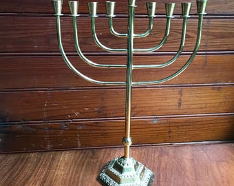 May include: A polished brass menorah featuring nine candle holders arranged in a symmetrical, curved design. The central candle holder is elevated above the others, supported by a slender vertical stem and a decorative, hexagonal base.