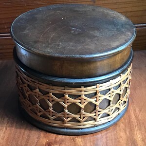 May include: A cylindrical wooden box with a dark brown finish and a matching lid. The base of the container is wrapped in a decorative, light-colored woven cane lattice pattern that contrasts with the smooth, dark wood of the lid and frame.