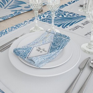 May include: A white dinner plate with a blue and white patterned napkin folded on top. The napkin has a diamond-shaped label with the text "Happy Passover" and "ל פסח שמח" in Hebrew. The plate is on a white tablecloth with a blue and white patterned runner.