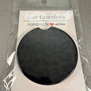 May include: A circular black car coaster featuring a subtle tonal leopard print pattern and a small notch at the top edge. The coaster is packaged in a clear plastic sleeve with a white backing card labeled Car Coasters Handmade with love.