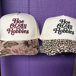 May include: Two off-white trucker hats featuring the embroidered text "Hoe Of All Hobbies" in a cursive font. The left hat has a floral tapestry-style brim, while the right hat features a black and tan leopard print pattern on the brim.
