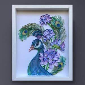 Op de afbeelding: Een ingelijst papier-quilling kunstwerk met een pauw met blauwe en groene veren en paarse bloemen. De kop van de pauw is naar de zijkant gedraaid en zijn oog is zichtbaar. Het kunstwerk is geplaatst tegen een witte achtergrond.