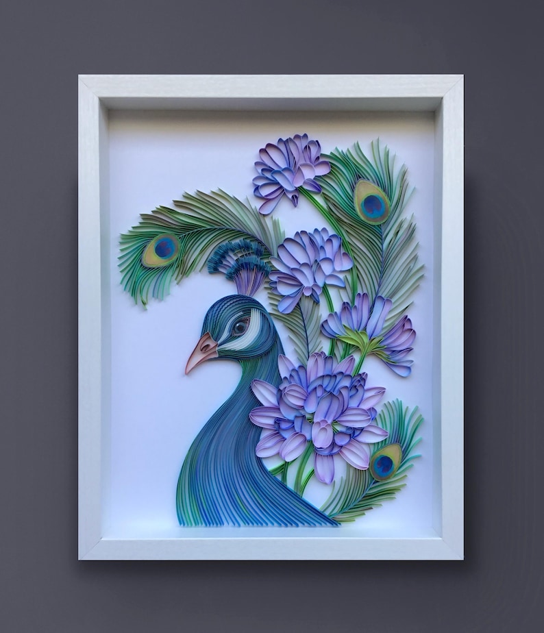 Op de afbeelding: Een ingelijst papier-quilling kunstwerk met een pauw met blauwe en groene veren en paarse bloemen. De kop van de pauw is naar de zijkant gedraaid en zijn oog is zichtbaar. Het kunstwerk is geplaatst tegen een witte achtergrond.
